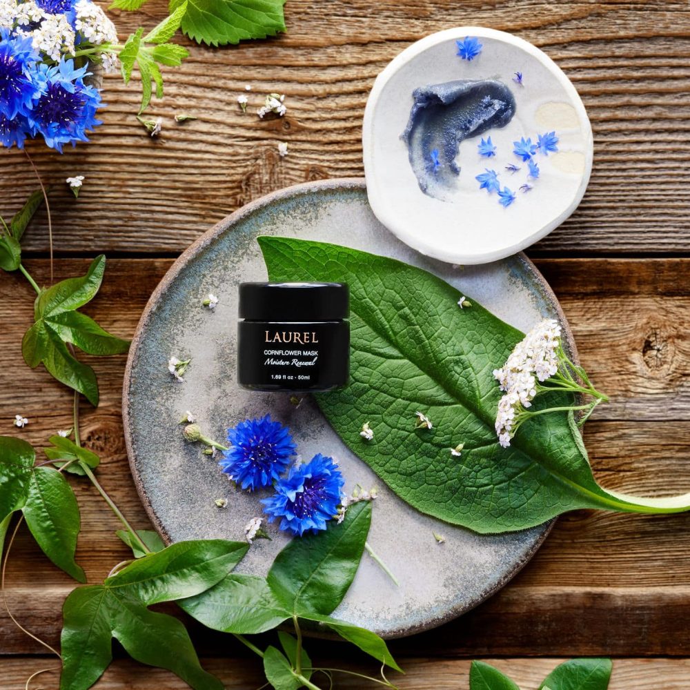 Jar or Cornflower Mask surrounded by green leaves and vibrant blue cornflower petals